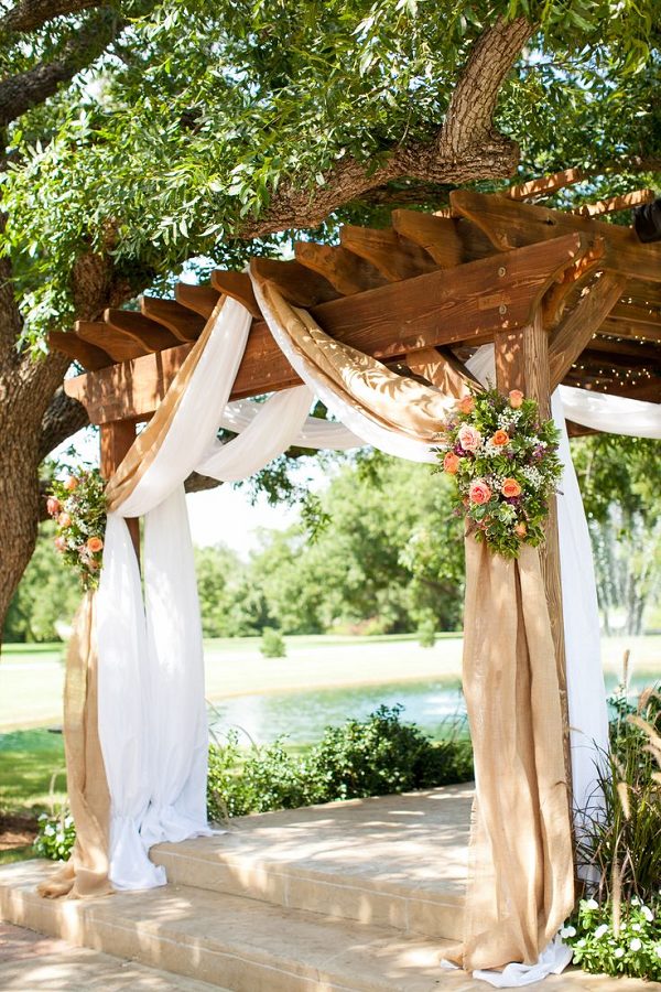 Burlap draping with country pink and green flowers over a wooden pergola
