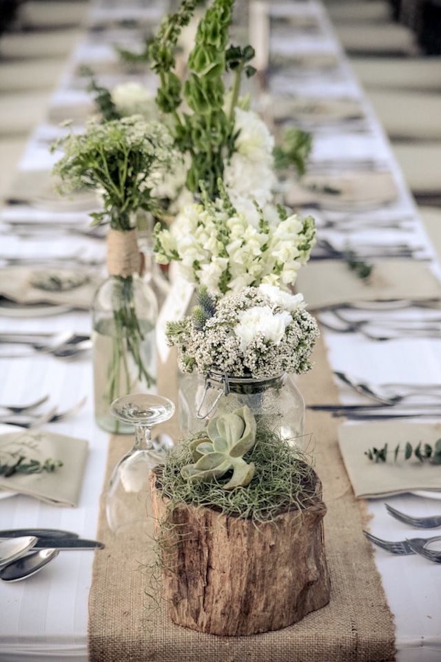Long table arrangement with a board running down the middle with mixed glassware, greenery, and candles. Greens, tans, and white table linens